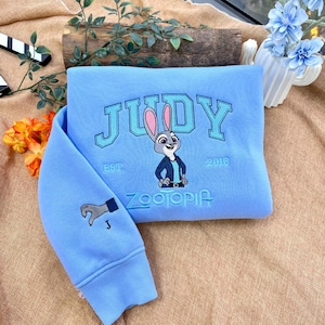 以下が含まれることがあります： 水色のスウェットシャツ。カレッジフォントで「JUDY」と刺繍され、ウサギの漫画の画像と「ZOOTOPIA」の文字が描かれています。袖には手のグラフィックがあります。「EST. 2016」もスウェットシャツに刺繍されています。