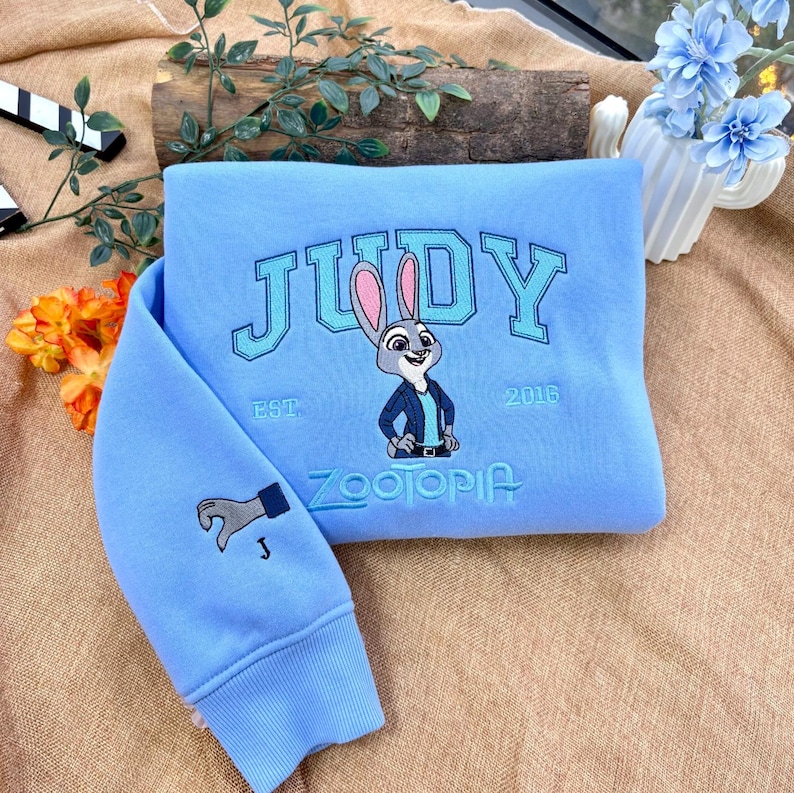 以下が含まれることがあります： 水色のスウェットシャツ。カレッジフォントで「JUDY」と刺繍され、ウサギの漫画の画像と「ZOOTOPIA」の文字が描かれています。袖には手のグラフィックがあります。「EST. 2016」もスウェットシャツに刺繍されています。