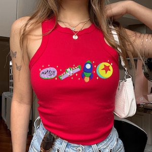 May include: A red vest top featuring embroidered cartoon graphics. The graphics include the word "ANDY", a space ship, a rocket, and a ball with a star. The top is sleeveless and made of a ribbed material.