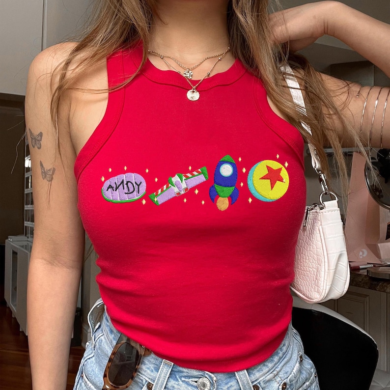May include: A red vest top featuring embroidered cartoon graphics. The graphics include the word "ANDY", a space ship, a rocket, and a ball with a star. The top is sleeveless and made of a ribbed material.