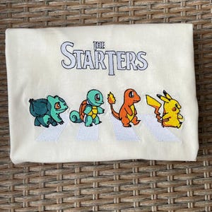 May include: Cream-colored t-shirt featuring an embroidered design of four cartoon characters walking across a crosswalk. The text "THE STARTERS" is embroidered above the characters. The characters are Bulbasaur, Squirtle, Charmander, and Pikachu.