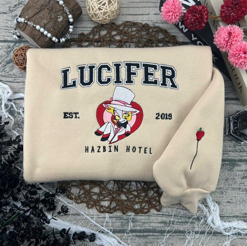 May include: Beige sweatshirt with the word "LUCIFER" in black collegiate lettering. Below is a cartoon character in a red heart, and the words "EST. 2019 HAZBIN HOTEL". The sleeve features a red apple embroidery.