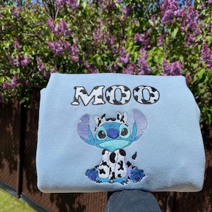 May include: A pale blue sweatshirt with a cartoon character design. The character is blue with large ears and a cow-like pattern. The word "MOO" is embroidered above the character in a black and white cow print.