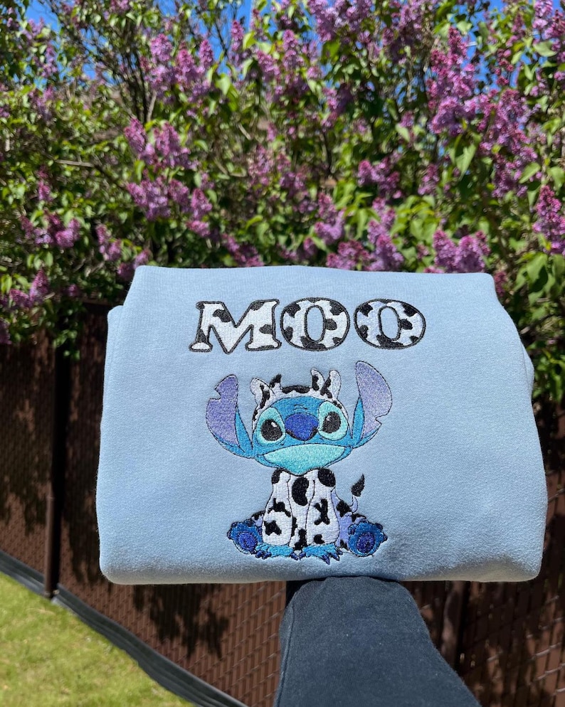 May include: A pale blue sweatshirt with a cartoon character design. The character is blue with large ears and a cow-like pattern. The word "MOO" is embroidered above the character in a black and white cow print.