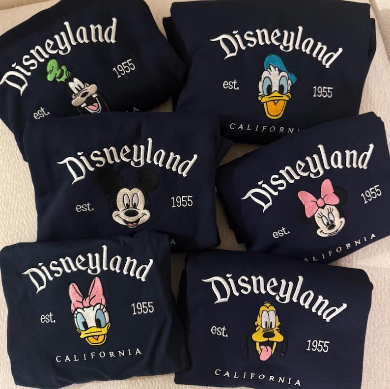 Peut inclure: Des sweat-shirts bleu marine avec des broderies "Disneyland" et des personnages. Chaque sweat-shirt pr&eacute;sente un personnage Disney diff&eacute;rent : Dingo, Donald Duck, Mickey Mouse, Minnie Mouse, Daisy Duck et Pluto. "est. 1955" et "CALIFORNIA" sont &eacute;galement brod&eacute;s.