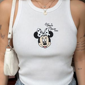 Może przedstawiać: Biały prążkowany top na ramiączkach z haftowanym wzorem Myszki Minnie. Wz&oacute;r zawiera twarz Myszki Minnie, kokardę w kropki oraz napis "Minnie Mouse xoxo" czarną czcionką. Top na ramiączkach jest bez rękaw&oacute;w.