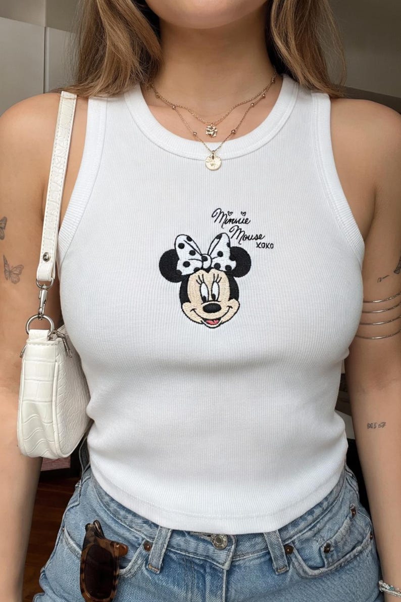 Może przedstawiać: Biały prążkowany top na ramiączkach z haftowanym wzorem Myszki Minnie. Wz&oacute;r zawiera twarz Myszki Minnie, kokardę w kropki oraz napis "Minnie Mouse xoxo" czarną czcionką. Top na ramiączkach jest bez rękaw&oacute;w.