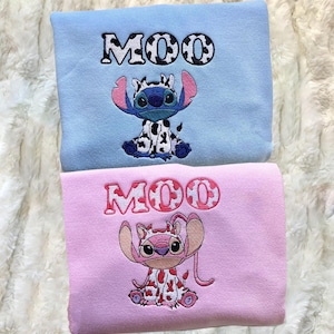 May include: A light blue sweatshirt featuring an embroidered blue character wearing a cow-print outfit with the text MOO above it, and a light pink sweatshirt featuring an embroidered pink character wearing a cow-print outfit with the text MOO above it.