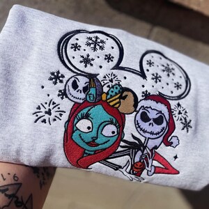 Puede incluir: Una sudadera gris claro con un dise&ntilde;o bordado de Sally y Jack Skellington de The Nightmare Before Christmas. El dise&ntilde;o incluye copos de nieve y orejas de Mickey Mouse. Sally tiene el pelo rojo y Jack lleva un gorro de Pap&aacute; Noel.