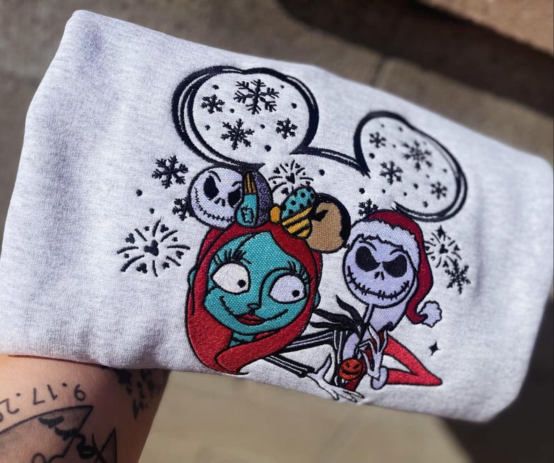Puede incluir: Una sudadera gris claro con un dise&ntilde;o bordado de Sally y Jack Skellington de The Nightmare Before Christmas. El dise&ntilde;o incluye copos de nieve y orejas de Mickey Mouse. Sally tiene el pelo rojo y Jack lleva un gorro de Pap&aacute; Noel.