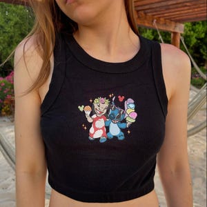 May include: Black ribbed tank top with a colorful embroidered graphic featuring two cartoon characters holding ice cream cones. The graphic includes a character in a red dress and a blue character, both with floral accents.