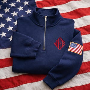 May include: A navy blue quarter-zip pullover sweatshirt featuring a silver-toned metal zipper. The chest displays a red embroidered monogram with three letters, and the left sleeve features an embroidered patch of the American flag.