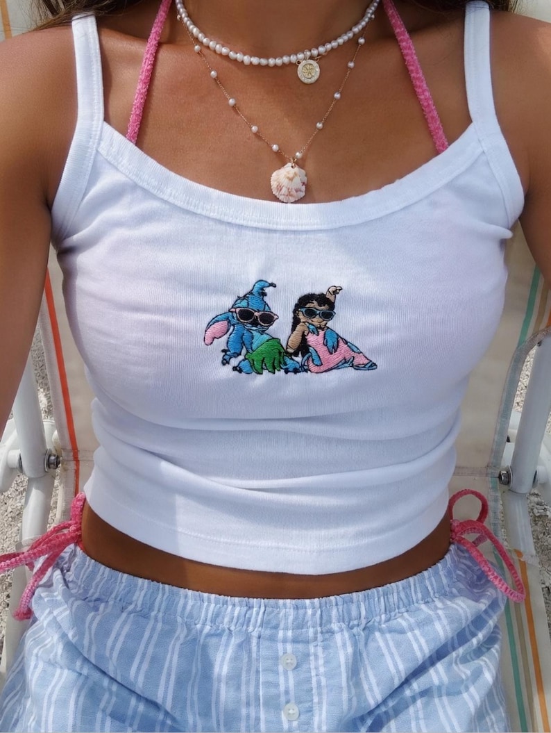 Puede incluir: Camiseta sin mangas blanca con un gr&aacute;fico bordado de dos personajes de dibujos animados con gafas de sol. La camiseta se combina con pantalones cortos a rayas azules y blancas. Un top de bikini de ganchillo rosa y collares de perlas completan el look.