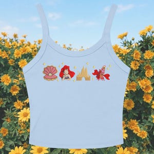 May include: Pale blue vest top featuring embroidered images of a seashell, Ariel, a castle, and Sebastian the crab. The top has thin straps. The background shows yellow flowers and a blue sky.