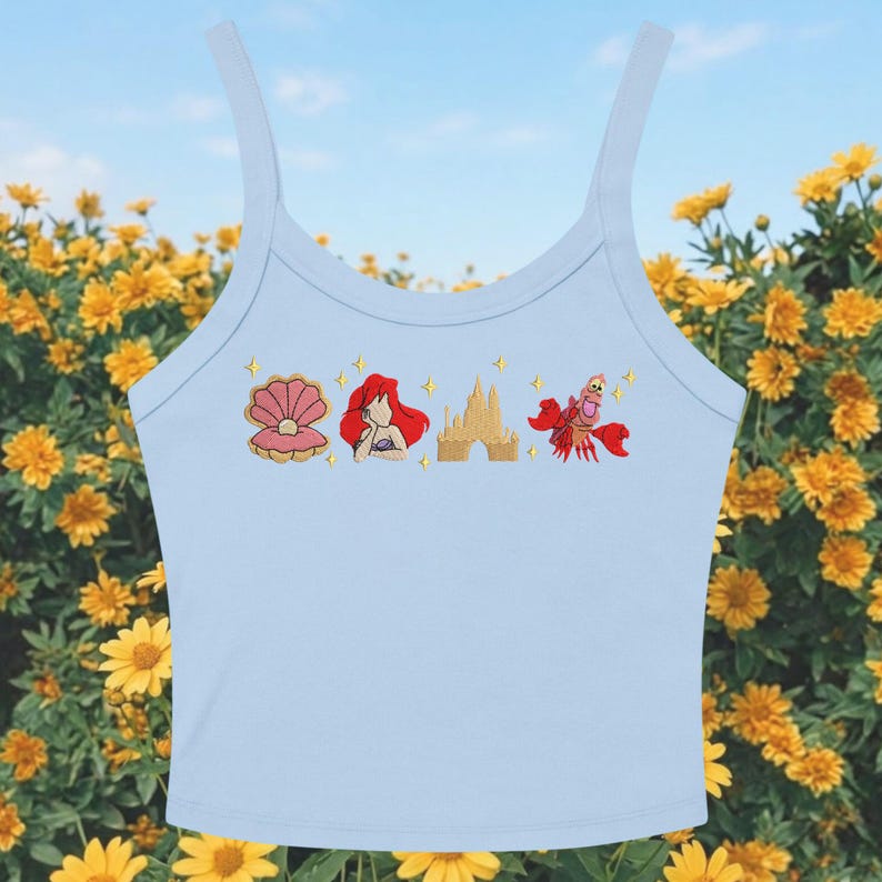 May include: Pale blue vest top featuring embroidered images of a seashell, Ariel, a castle, and Sebastian the crab. The top has thin straps. The background shows yellow flowers and a blue sky.