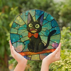 May include: Round stained glass art piece featuring a black cat with a red bow tie. The cat has large yellow eyes and is set against a colorful stained glass background with shades of blue, green, and yellow. The art piece is held up by hands.