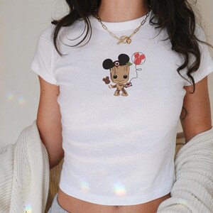 May include: White crop top featuring a cartoon character Groot with Mickey Mouse ears and a red balloon. The graphic is in the center of the shirt. The shirt is short-sleeved and has a ribbed texture.