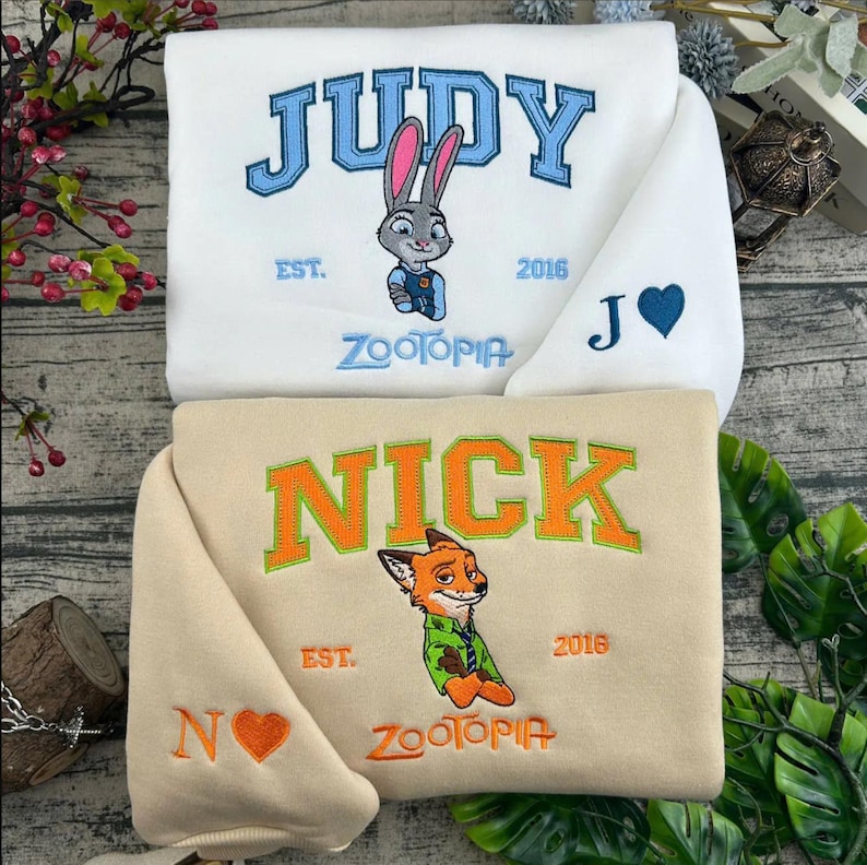 Puede incluir: Dos sudaderas, una blanca y otra beige, con dise&ntilde;os bordados. La sudadera blanca tiene "JUDY" en azul, un gr&aacute;fico de conejo y el texto "ZOOTOPIA". La sudadera beige dice "NICK" en naranja, un gr&aacute;fico de zorro y el texto "ZOOTOPIA".