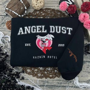 May include: Black sweatshirt with white embroidered text "ANGEL DUST" above a pink and white heart-shaped graphic. The graphic features a cartoon character with a red bow tie. The text "EST. 2019" and "HAZBIN HOTEL" are also visible.
