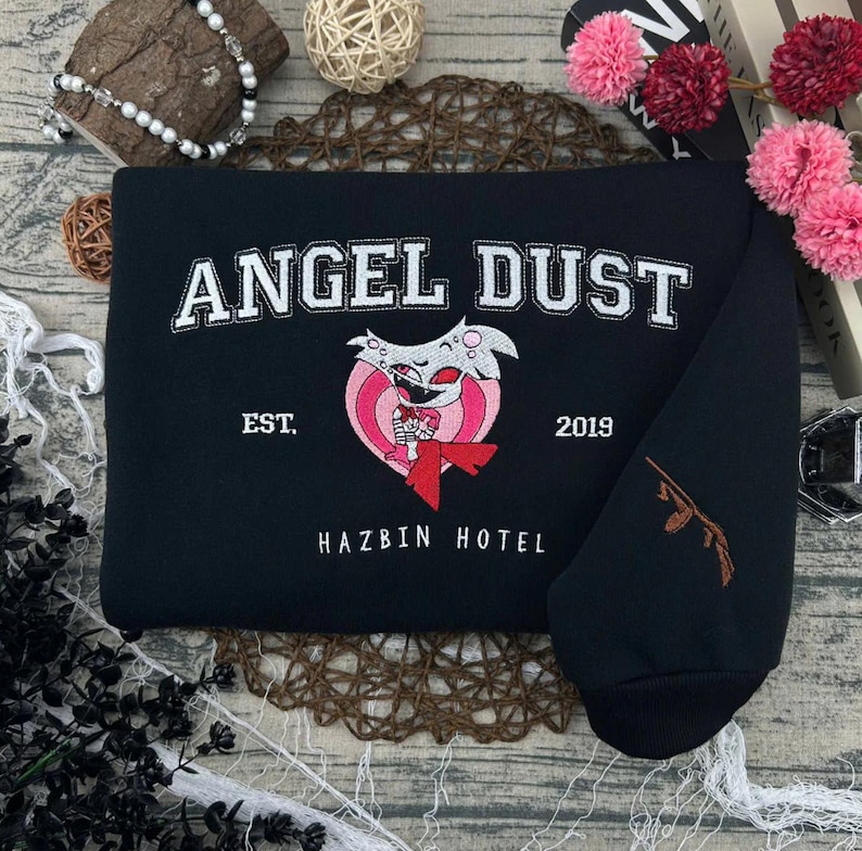 May include: Black sweatshirt with white embroidered text "ANGEL DUST" above a pink and white heart-shaped graphic. The graphic features a cartoon character with a red bow tie. The text "EST. 2019" and "HAZBIN HOTEL" are also visible.