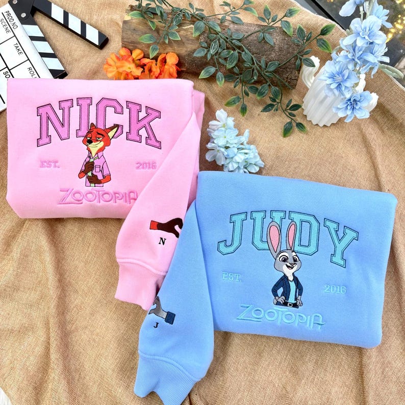 以下が含まれることがあります： 刺繍デザインが施されたピンクとブルーのスウェットシャツ2枚。ピンクのスウェットシャツにはキツネのグラフィックと「NICK」、ブルーにはウサギのグラフィックと「JUDY」の文字。どちらも「ZOOTOPIA」と「2016」または「2018」の文字入り。