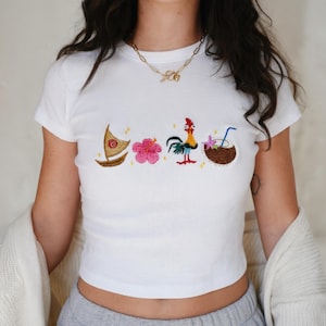 May include: White t-shirt with embroidered designs of a boat, pink flower, rooster, and coconut drink. The shirt has short sleeves and a crew neck. The design is colorful and detailed, with a tropical theme.