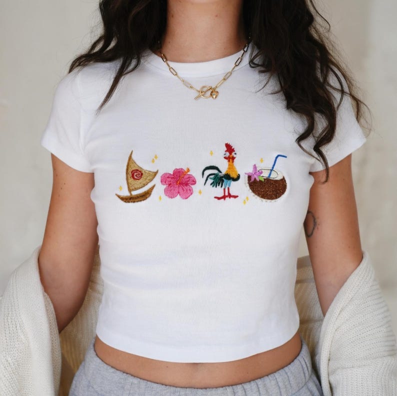 May include: White t-shirt with embroidered designs of a boat, pink flower, rooster, and coconut drink. The shirt has short sleeves and a crew neck. The design is colorful and detailed, with a tropical theme.