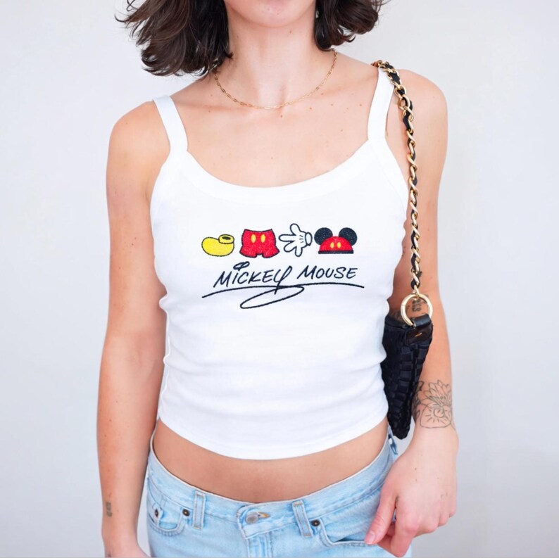 May include: White tank top with a Mickey Mouse graphic. The graphic features a yellow shoe, red shorts, a white glove, and Mickey's red and black ears. The words "Mickey Mouse" are written in black script below the graphic.