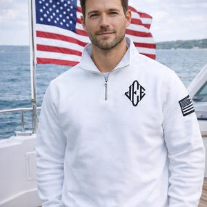 Könnte beinhalten: Weißes Sweatshirt mit Viertelreißverschluss, schwarzem Monogramm und einem amerikanischen Flaggenaufnäher am Ärmel. Das Sweatshirt hat einen Stehkragen und einen silbernen Reißverschluss. Der Hintergrund zeigt eine amerikanische Flagge.