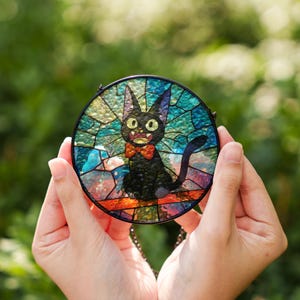 May include: A round stained glass piece featuring a black cat with a red bow tie. The cat has large, yellow eyes and is set against a colorful background of blue, green, and orange glass pieces. The piece is held by hands.