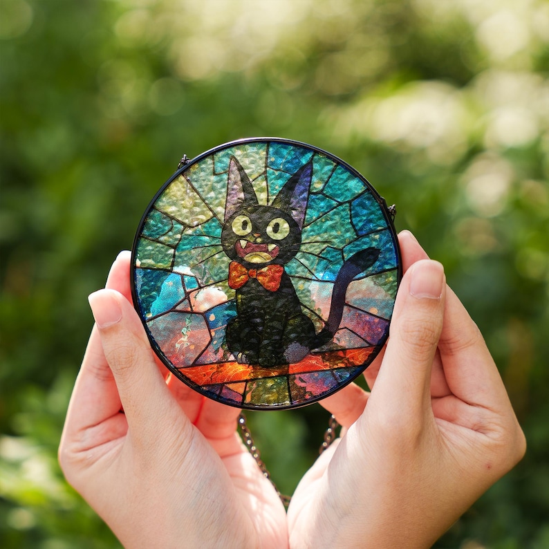 May include: A round stained glass piece featuring a black cat with a red bow tie. The cat has large, yellow eyes and is set against a colorful background of blue, green, and orange glass pieces. The piece is held by hands.