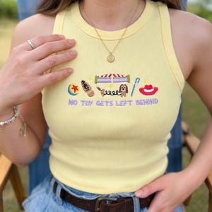 May include: Yellow ribbed tank top with embroidered cartoon graphics and the text "NO TOY GETS LEFT BEHIND". The design includes a ball, a boot, a circus tent, a dog, and a cowboy hat.