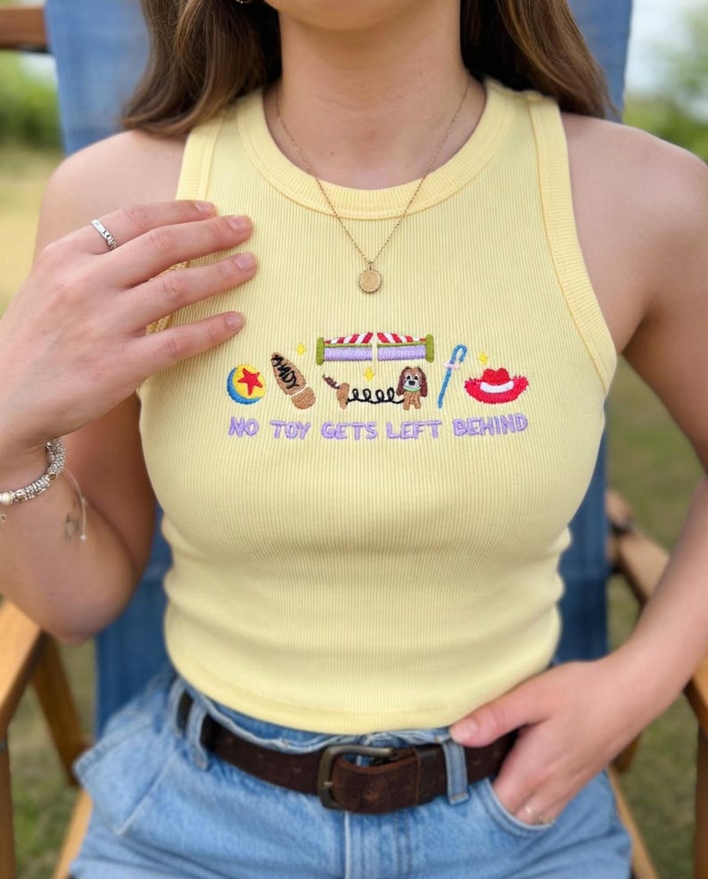 May include: Yellow ribbed tank top with embroidered cartoon graphics and the text "NO TOY GETS LEFT BEHIND". The design includes a ball, a boot, a circus tent, a dog, and a cowboy hat.