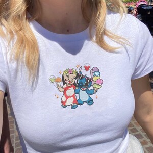 May include: White t-shirt featuring a colorful embroidered design of two cartoon characters holding ice cream cones. One character is wearing a floral crown and a red dress, while the other is blue. The shirt has a crew neck and short sleeves.