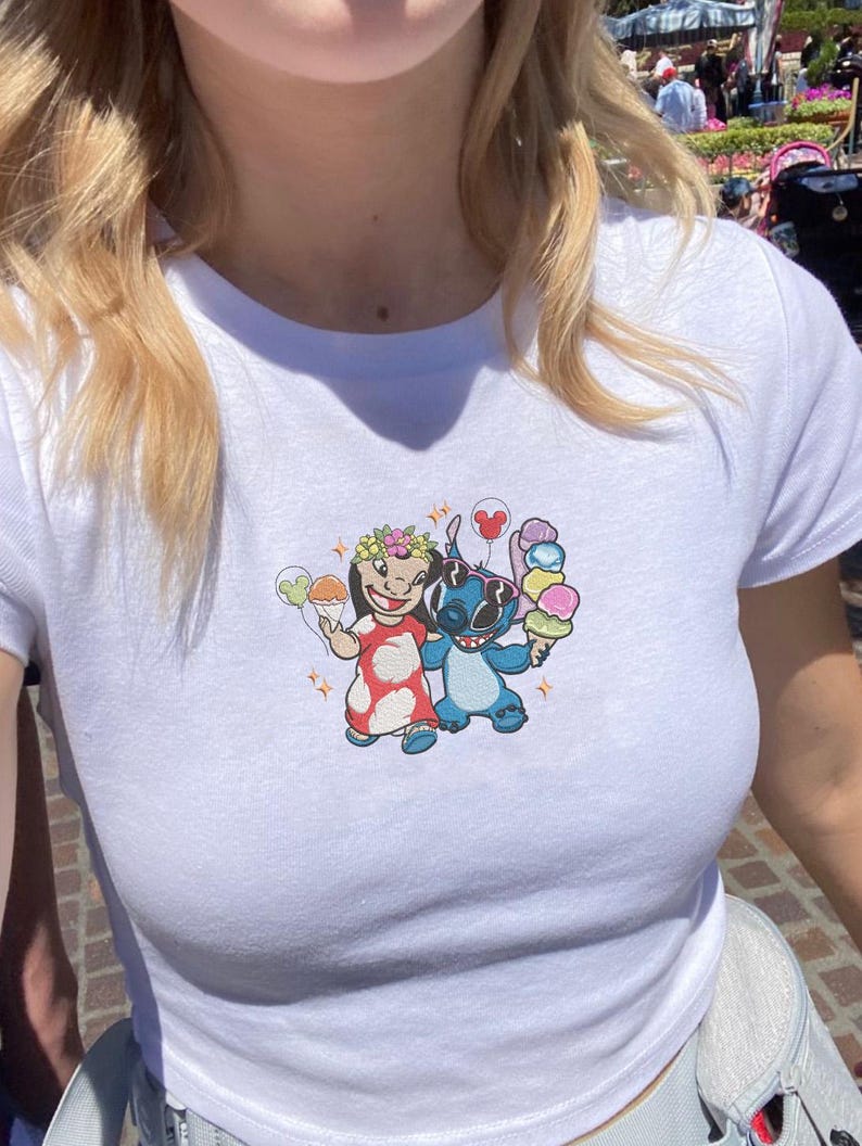 May include: White t-shirt featuring a colorful embroidered design of two cartoon characters holding ice cream cones. One character is wearing a floral crown and a red dress, while the other is blue. The shirt has a crew neck and short sleeves.