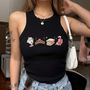 Può includere: Una canotta nera a coste con un ricamo raffigurante una tazzina, un diadema dorato con una rosa rosa, una pila di libri e una piccola creatura. Il top è abbinato a jeans in denim azzurro e a una borsa a spalla bianca.