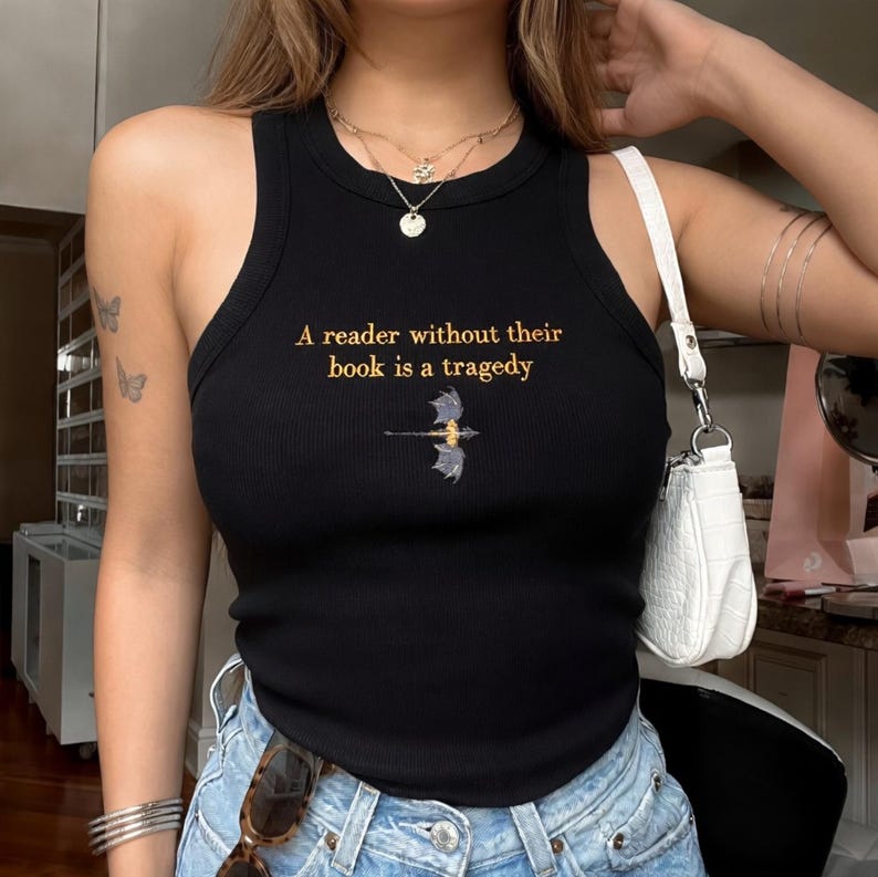 Puede incluir: Camiseta sin mangas negra acanalada con el texto dorado "A reader without their book is a tragedy". Un peque&ntilde;o gr&aacute;fico de drag&oacute;n est&aacute; debajo del texto. La camiseta es sin mangas y tiene cuello redondo.