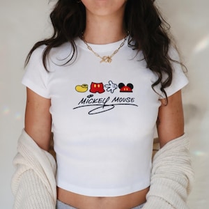 May include: White ribbed crop top featuring a Mickey Mouse design. The design includes a yellow shoe, red shorts, a white glove, and Mickey's face. The words "Mickey Mouse" are written in black script below the design.