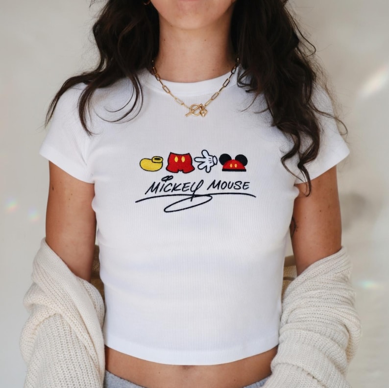 May include: White ribbed crop top featuring a Mickey Mouse design. The design includes a yellow shoe, red shorts, a white glove, and Mickey's face. The words "Mickey Mouse" are written in black script below the design.