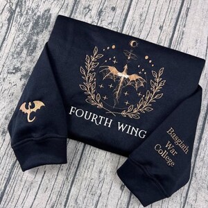 May include: Black sweatshirt with gold dragon and text. The front displays a dragon design within a laurel wreath and the words "FOURTH WING." The left sleeve has a dragon emblem, and the right sleeve says "Basgiath War College."