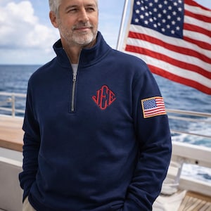 May include: A navy blue quarter-zip pullover featuring a red embroidered monogram on the chest and a small American flag patch on the left sleeve. The garment has a high collar with a silver-toned zipper and long sleeves with ribbed cuffs.