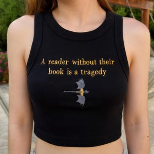 Puede incluir: Camiseta sin mangas negra acanalada con la cita "A reader without their book is a tragedy" en letras doradas. Un gr&aacute;fico de drag&oacute;n dorado y azul est&aacute; centrado debajo del texto. La camiseta sin mangas es sin mangas.