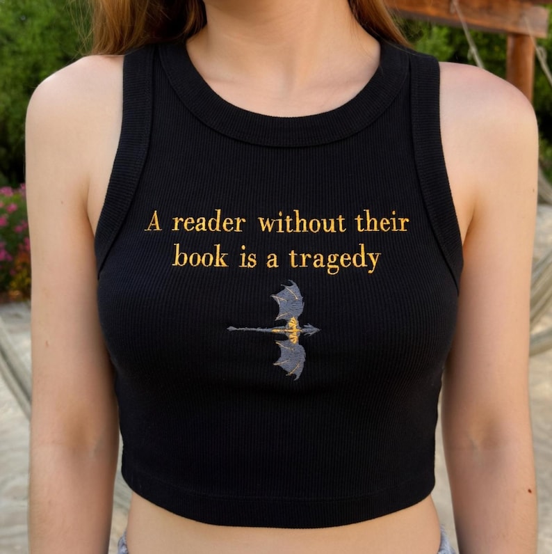 Puede incluir: Camiseta sin mangas negra acanalada con la cita "A reader without their book is a tragedy" en letras doradas. Un gr&aacute;fico de drag&oacute;n dorado y azul est&aacute; centrado debajo del texto. La camiseta sin mangas es sin mangas.