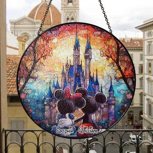 Op de afbeelding: Ronde glas-in-loodraam met een kleurrijke kasteelscène met Mickey en Minnie Mouse. Het kunstwerk heeft een zwart metalen frame en een ketting om op te hangen. De namen "Sophia William" staan onderaan.