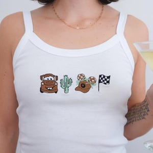 May include: A white tank top featuring a horizontal row of four embroidered designs: a brown tow truck, a light green cactus, a brown baseball cap with mouse ears and a light green bow, and a black and white checkered racing flag.