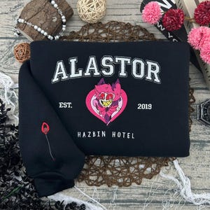 May include: Black sweatshirt with the word "ALASTOR" in white block letters. Below the word is "EST. 2019" and "HAZBIN HOTEL". A pink heart with a cartoon character is in the center. A red and black design is on the sleeve.
