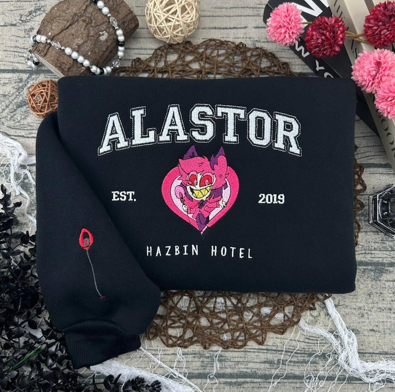 May include: Black sweatshirt with the word "ALASTOR" in white block letters. Below the word is "EST. 2019" and "HAZBIN HOTEL". A pink heart with a cartoon character is in the center. A red and black design is on the sleeve.