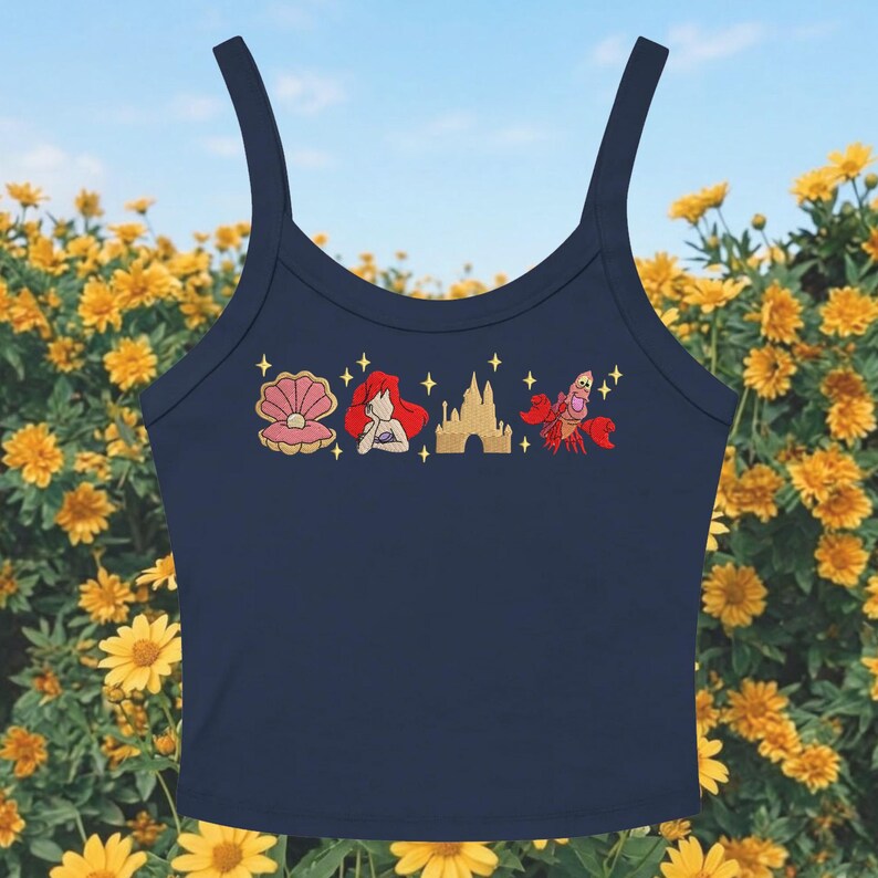 May include: A navy blue crop top featuring embroidered images of a seashell, a red-haired mermaid, a castle, and a red crab. The top has thin straps and is set against a backdrop of yellow flowers and a blue sky.