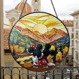 Op de afbeelding: Ronde glas-in-lood raam met Mickey en Minnie Mouse voor een berglandschap met zonsondergang. Het raam is zwart omlijst en hangt aan een ketting. De namen "Rose" en "Jack" staan onderaan.