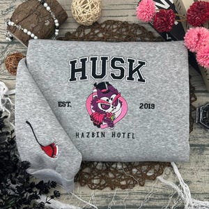 May include: A gray sweatshirt with the word "HUSK" in black block letters above a cartoon character in a pink circle. The sleeve features a red and black broom design. The text "EST. 2019" and "HAZBIN HOTEL" are also visible.